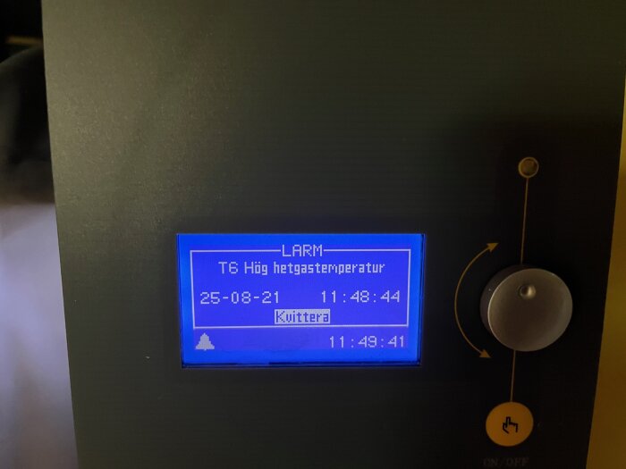 Visar display på värmepump med larmmeddelande "Hög hetgastemperatur", datum och tid samt en knapp för att kvittera larmet.