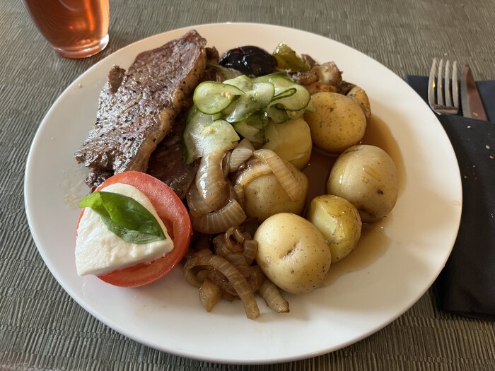 Tallrik med ryggbiff, stekt lök, nykokt potatis, inlagd gurka, sås och en tomatskiva med mozzarella och basilika.