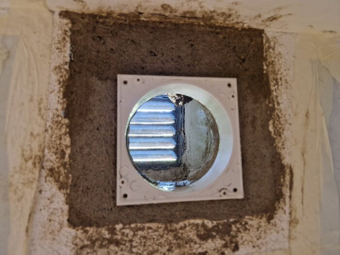 Ventilöppning med cementsockel och monterad ram för innerdel. Bakom öppningen syns ljuset genom ventilgallret.