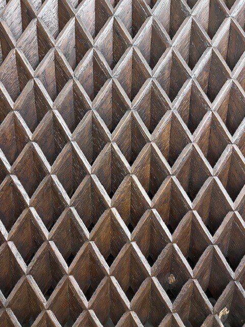 Ventilationsgaller i mörkt trä med ett geometriskt mönster av pyramidformade strukturer som bildar ett rutmönster.