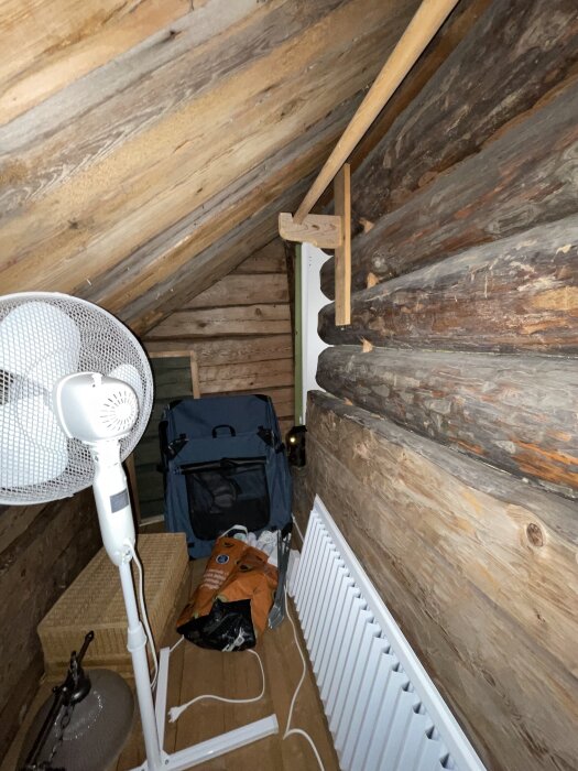 En smal vind med träpaneler, en golvfläkt, en korg, en väska och diverse föremål, samt synlig del av ett element.