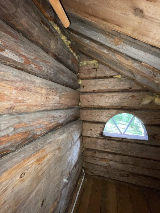 Trätak och timmerväggar med ett halvmåneformat fönster i ett rum, synlig isolering i hörnen.