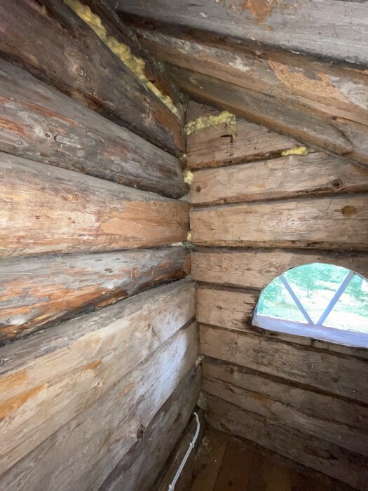Vägg i trä med synliga stockar och isolering, ett fönster med båge syns till höger.