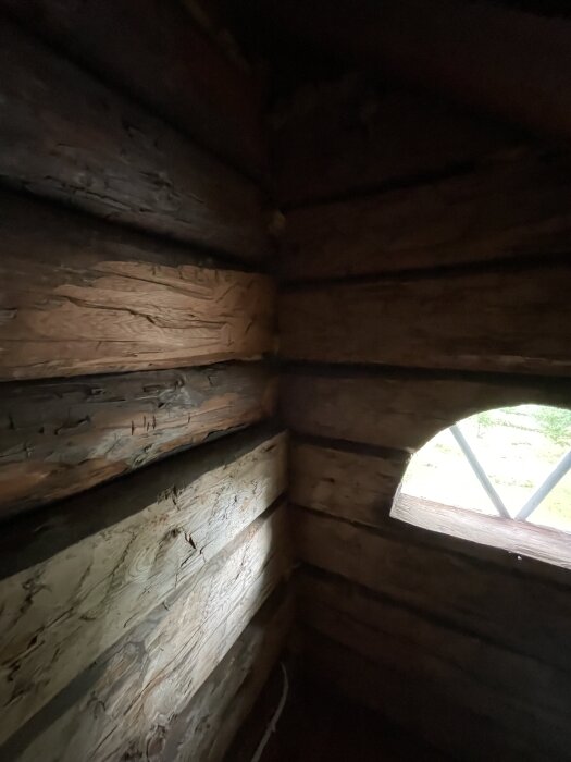 En insida av en vägguppbyggnad med timmerstockar, belyst av svagt ljus. En rundad öppning med en vy utanför syns till höger.