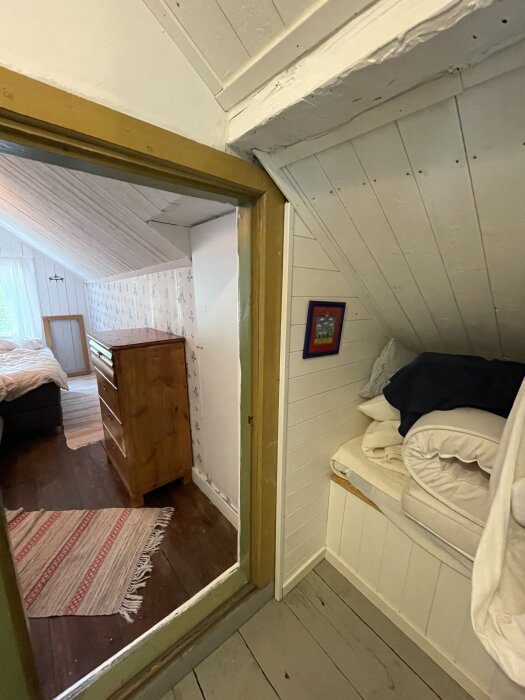 En snedtaksgarderob i ett rum med träväggar och -golv, anslutande till ett sovrum med säng och byrå.