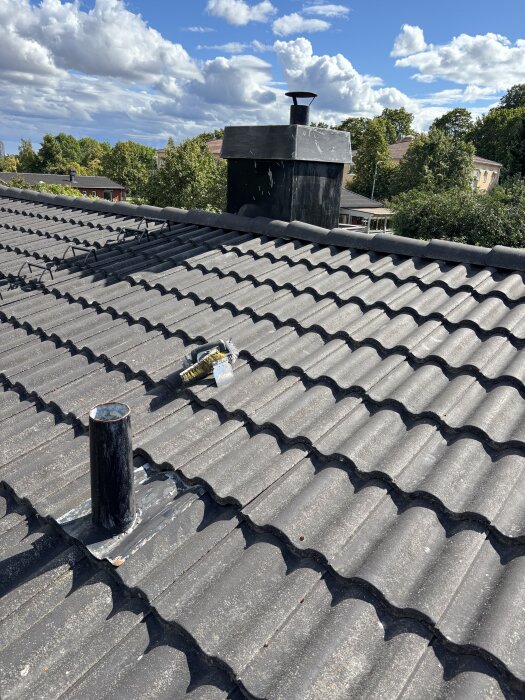 Skorsten och rör utan lock på ett 70-tals villatak under klarblå himmel med träd i bakgrunden.