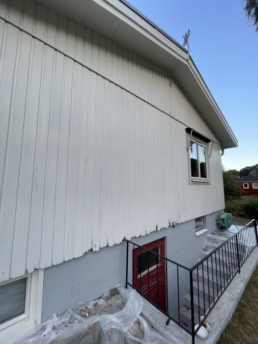 Vit träfasad med skadade, murkna plankor längst ner ovanför en röd dörr och grå betong grund. Träet visar tecken på försämring.