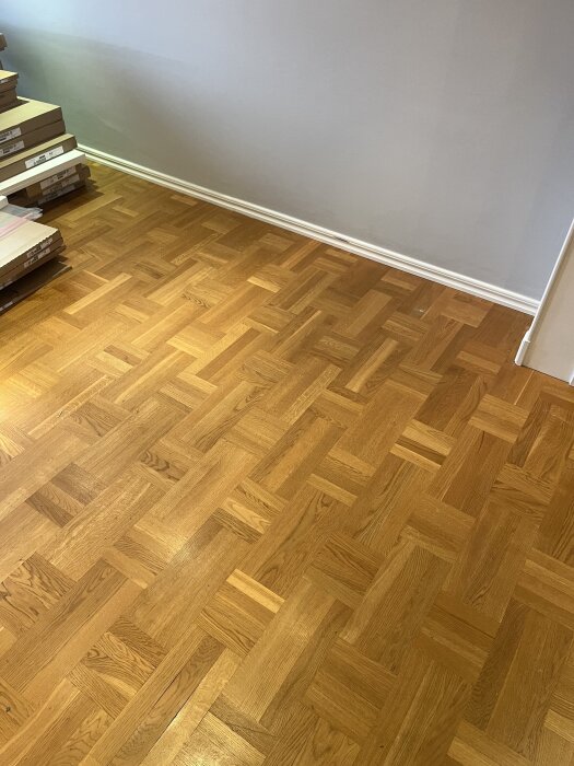 Golv av holländsk parkett med rutmönster i olika stavstorlekar, liknande 330x165 och 165x165 mm, med en stapel golvpaket vid väggen.