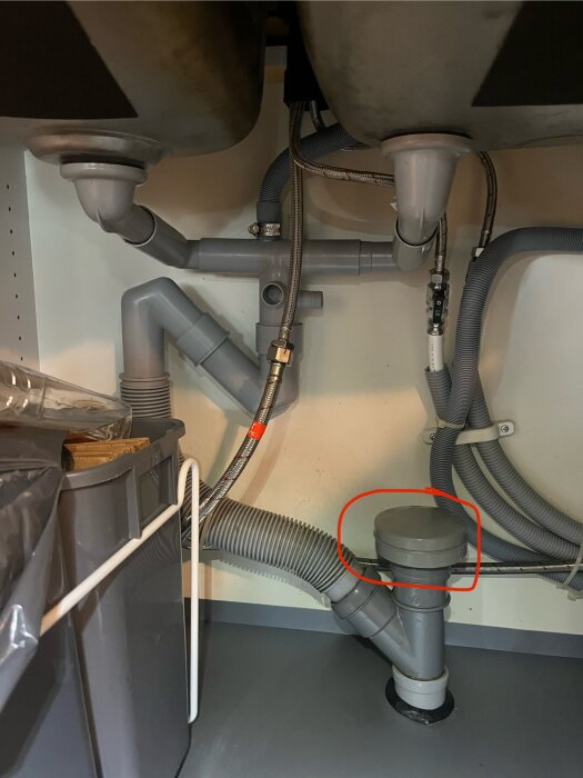 Under diskbänk med avloppsrör och vattenlås, där ett markerat lock på sidan av utloppet verkar vara ihåligt och orsaka luktproblem.