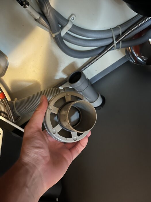 En människa håller i ett grått, ihåligt avloppslock i en miljö med rör och kablar under diskbänk.