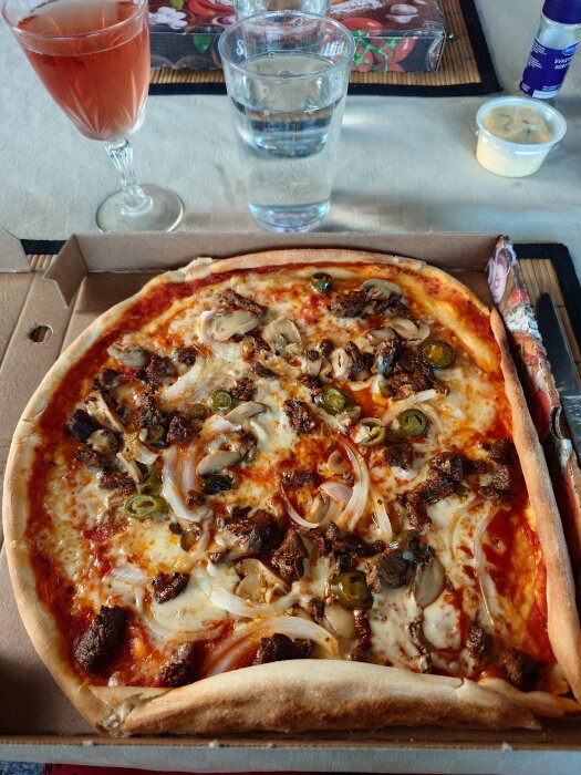 Pizza Acapulco med kött, svamp och jalapeño i en kartong, serverad med ett glas rosé och vatten på ett bord.