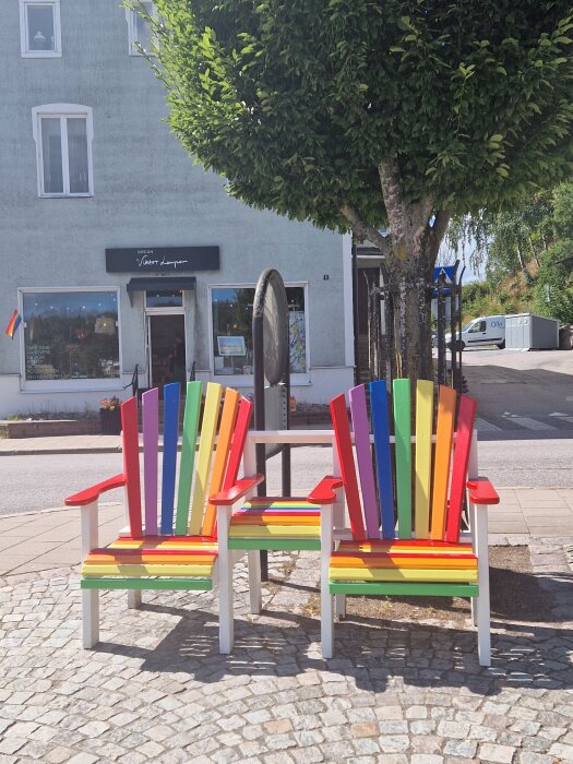 Två regnbågsfärgade adirondack-stolar på en stenlagd trottoar, bredvid ett träd framför en byggnad med en butiksskylt.