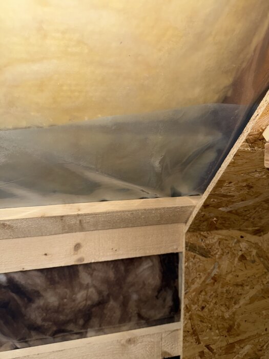 Bild av en snedtaksvägg med synlig brun isolering och träreglar, eventuellt i ett område bakom en byrå nära kattvinden.