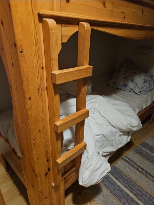 Trästege monterad på väggen intill platsbyggda våningssängar i en fjällstuga, med vit sängkläder och randig matta i träinredning.