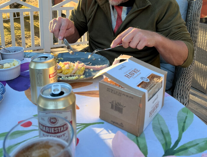 En person Àter mat utomhus vid ett bord med Mariestads ölburkar, smör, kexförpackning och en mÄltid med rÀkor.