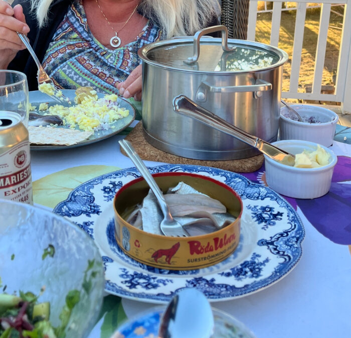 Matbord med surströmming, potatis och bröd samt en kastrull och smör. En Mariestads ölburk stÄr vid sidan av.