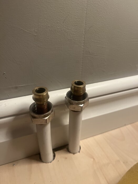 Två rör med metallkopplingar som sticker upp från golvet nära en vit golvlist.