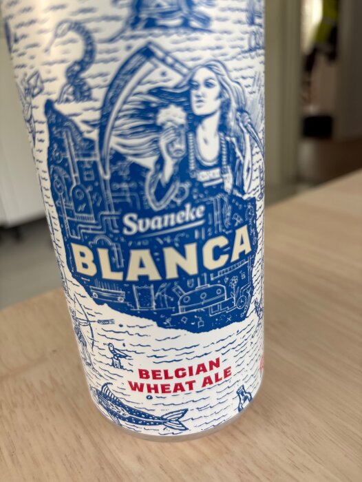 Vit burk med blå illustrationer innehållande Svaneke Blanca Belgian Wheat Ale, stående på ett bord.