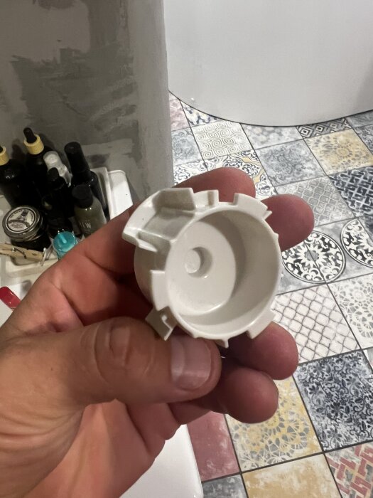 En hand håller en vit reservdel till spolknapp framför ett golv med färgglada kakelplattor.