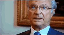 Jag kan inte identifiera personen på bilden, men bilden visar en äldre man som ser skeptisk ut i ett formellt rum.