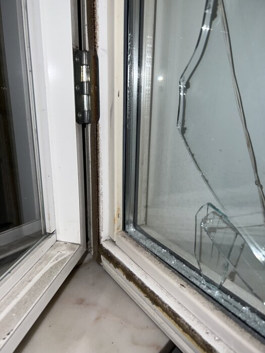 Trasigt fönster med sprucken glasruta och rostig gångjärnssida, nära detalj av fönsterkarm och öppna fönsterbågar.