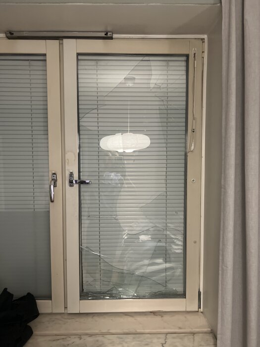 Sprucken glasruta i ett fönster med vita persienner, placerad i en träram med ett handtag och skruv synlig ovanpå.