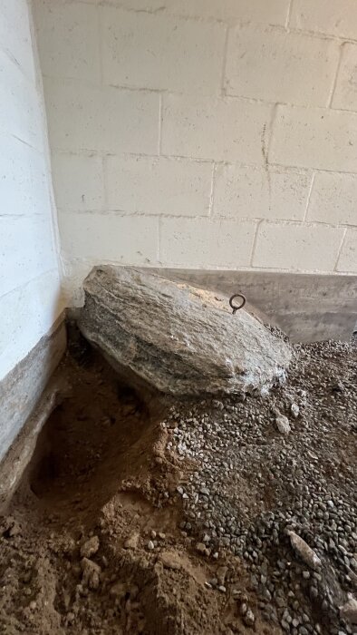 Stort stenblock i källarväggen med betongmur och jord, ögla fastsatt i stenen för eventuell borttagning.