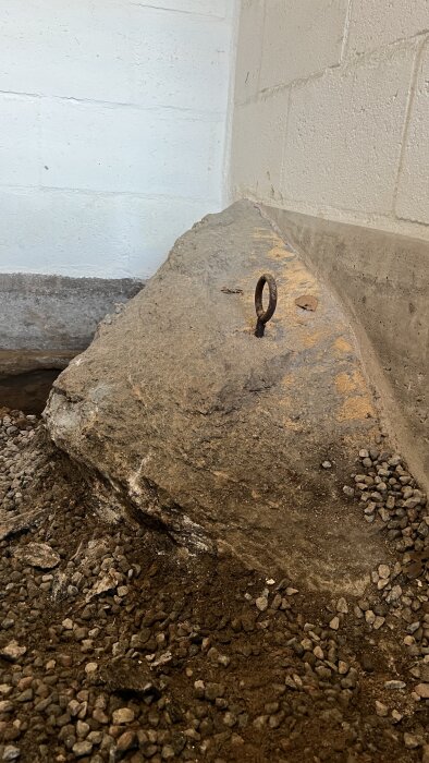 En stor sten i ett källarutrymme med en ögla på toppen, omgiven av grus, intill en murad vägg.