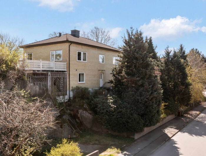 Tegelhuset med en altan och tät vegetation, bredvid en gata. Möjligt projektområde för altan och parkering.