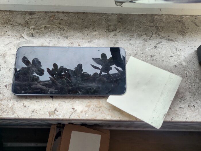 En mobiltelefon med reflekterande skärm ligger på ett fönsterbräde bredvid ett tomt papper.