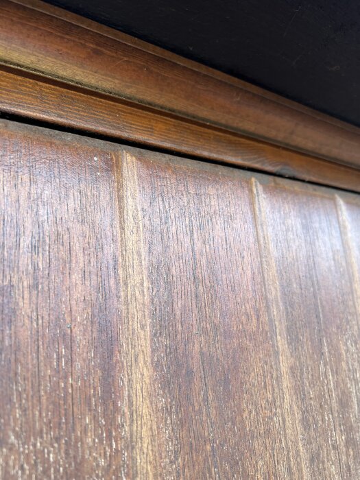 Äldre träpanel på ytterdörr med jämn slityta, potentiellt från 70-talet, som tidigare oljats men ej slipats, närbild av dörrens övre del.