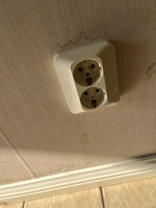 Fuktfläckar på vägg nära ett dubbeluttag i ett kök eller hall, potentiella tecken på mögel eller vattenskada.