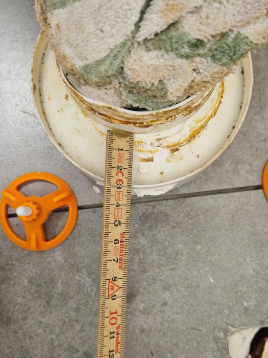 Gammalt toalettavlopp med brunt tätningsmaterial och måttband liggande på grått golv, två orange verktyg syns bredvid.