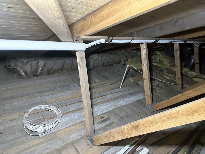 Loft med ventilationsrör, träreglar och isolering synliga; kablar ligger på golvet.