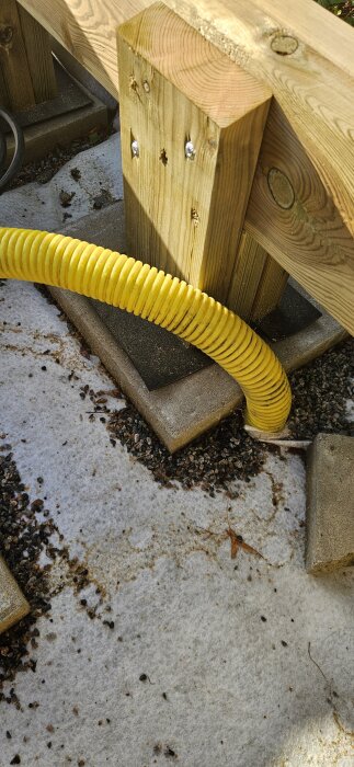 Gul korrugerad kabel som löper under en träkonstruktion på betongplattor, omgiven av småsten.