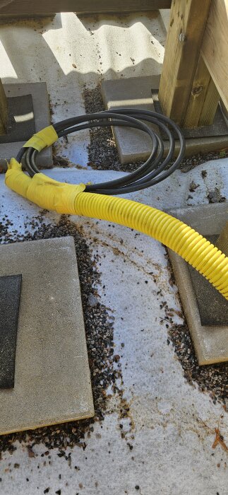 Elektriska kablar och gul skyddsslang ligger på en betongyta med grus, bredvid träkonstruktion på uteplats.