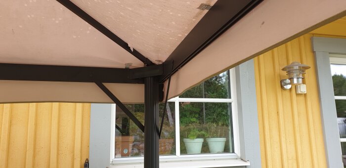 Pergolatak i närbild med metallstruktur, fäst vid en gul träfasad med ett fönster och lampa i bakgrunden.