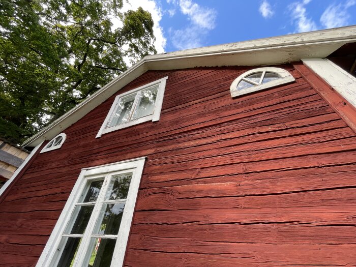 Röd trähusgavel med vita fönster och synliga stockar under en blå himmel.