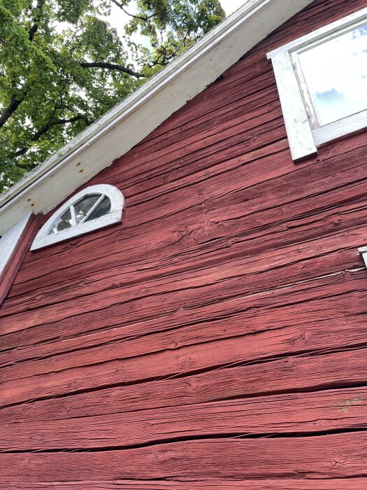 Röd träfasad på en husgavel med vita fönsterlister och en bågformad öppning under taket, omgiven av grönska.