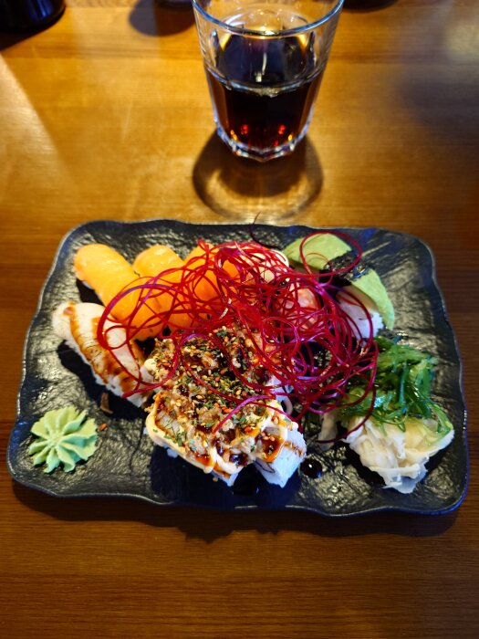 Sushitallrik med lax, röd trÄdliknande dekoration, wasabi och ett glas mörk dryck pÄ ett bord pÄ Dala Sushi i BorlÀnge.