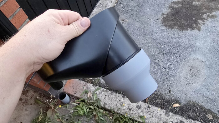Handen håller en svart och grå lövavskiljare till stuprör mot garaget, nyinstallerad med anpassad 3D-printad adapter.