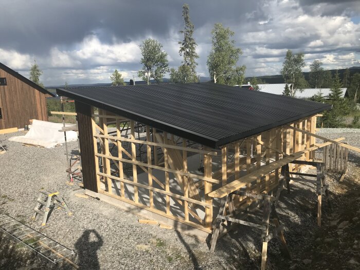 Bygge av en träram för en byggnad med tak, omgiven av träd och grus. Stegar och byggmaterial syns i förgrunden.