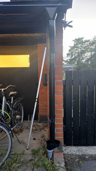 Svart stuprör monterat mot en röd tegelvägg intill en cykel, med en lövavskiljare och en grå adapter vid markröret.
