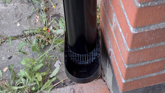 Svart stuprör monterat vid röd tegelvägg, sett från sidan, med en lövavskiljare vid röröppningen. Omgivande grönska på betongplattor.