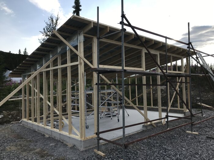 Träram för ett bygge med takstolar och byggnadsställningar på grusbelagd grund, omgiven av träd och himmel.