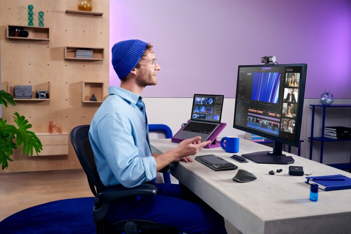 En person på hemmakontor använder en laptop och en stor monitor med Logitech tangentbord, mus och webbkamera. Modern arbetsplats med ergonomiska tillbehör.