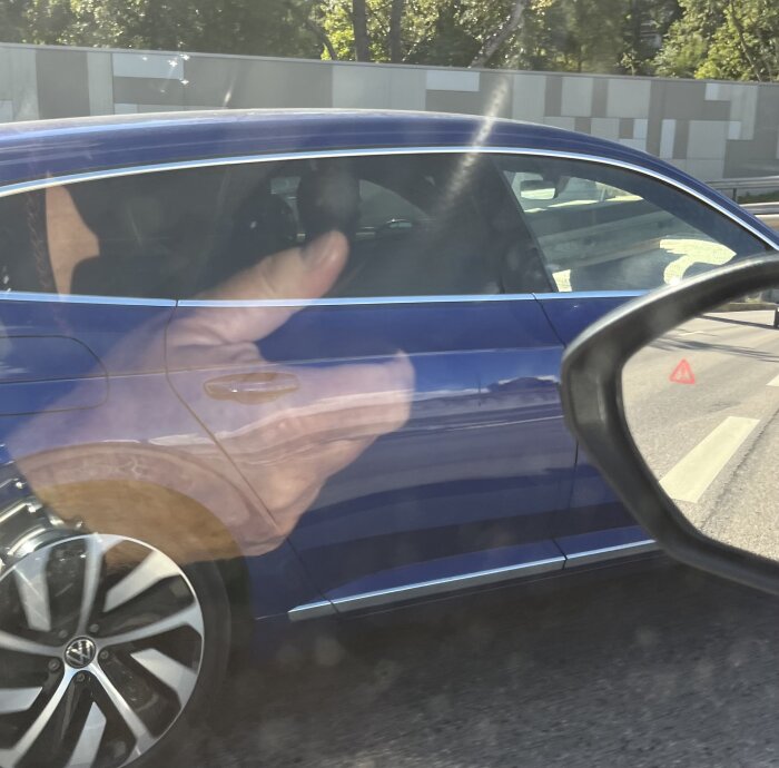 Blå bil med långa träreglar synliga genom bakrutan, reflektion av hand ger tummen upp i förgrunden.
