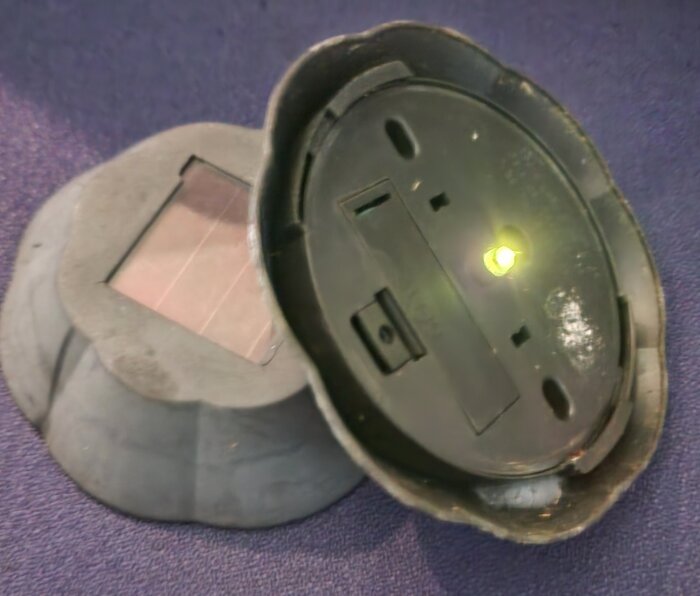 Solcellsdriven trädgårdslampa med öppen undersida, där en liten lampa fortfarande lyser svagt, visar slitage och gulnad yta.