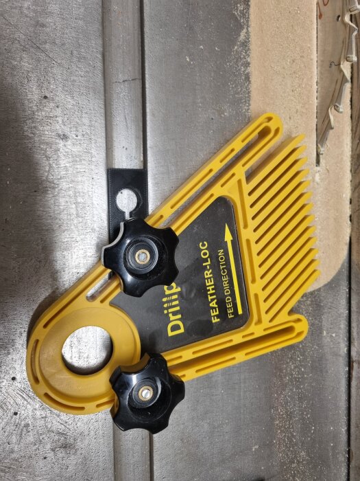 Gult fjäderanhåll monterat på en arbetsyta, med svarta vred och texten "DRiiiP FEATHER-LOC FEED DIRECTION" synlig.