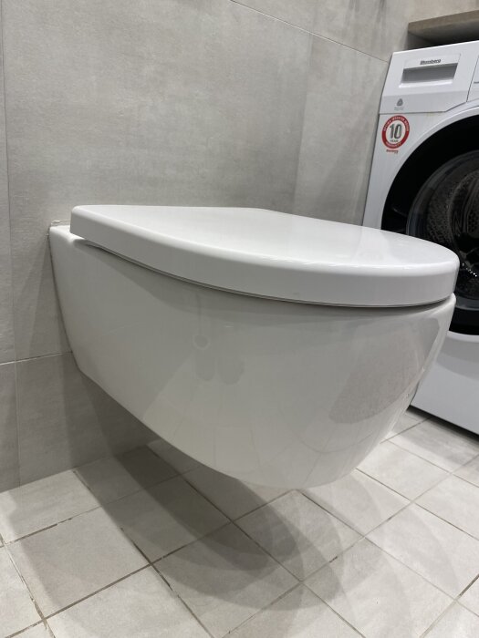 Vägghängd toalett med stängt lock, monterad i ett kaklat badrum. En tvättmaskin syns i bakgrunden. Toalettsitsen är trasig i fästet.